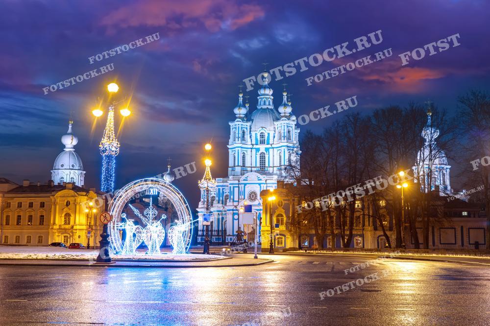 На фотографии изображен Смольный собор в Санкт-Петербурге вечером. Фасады украшены новогодней иллюминацией, создающей праздничное настроение. Зимний вечер подчеркивает красоту архитектуры собора и окружающих зданий. Световые инсталляции на площади перед собором добавляют волшебства.